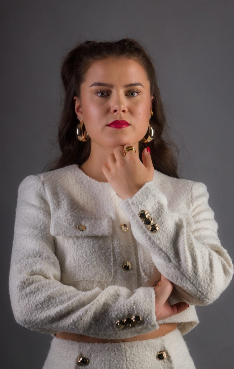 LIIVIA mit gelockten Haaren und rotem Lippenstift, in weißer Jacke mit Goldschmuck, selbstbewusst.