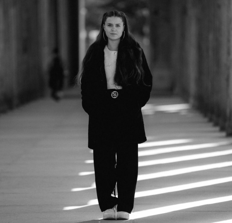 Junge Frau in schwarzem Outfit steht allein in einem lichtdurchfluteten Gang.